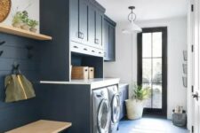 a stylish navy laundry mudroom with shaker cabinets, a washing machine and a dryer, a built-in bench and an open shelf