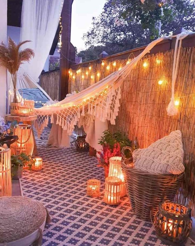 a boho balcony with an embroidered rug, lots of candle lanterns, a hammock with pillows, lamps and pillows and a jute pouf