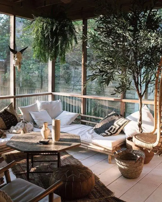 a boho screened porch with a corner rattan sofa with pillows, a unique Eastern coffee table, a leather pouf, potted plants