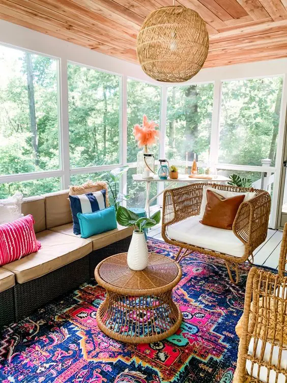 a bright boho screened porch with a dark wicker sofa and bold pillows, rattan chairs and a table, a colorful rug and a console table with candle lanterns