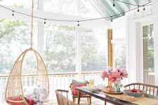 a bright screened porch with a suspended chair, a stained table, benches and chairs, strign lights and greenery