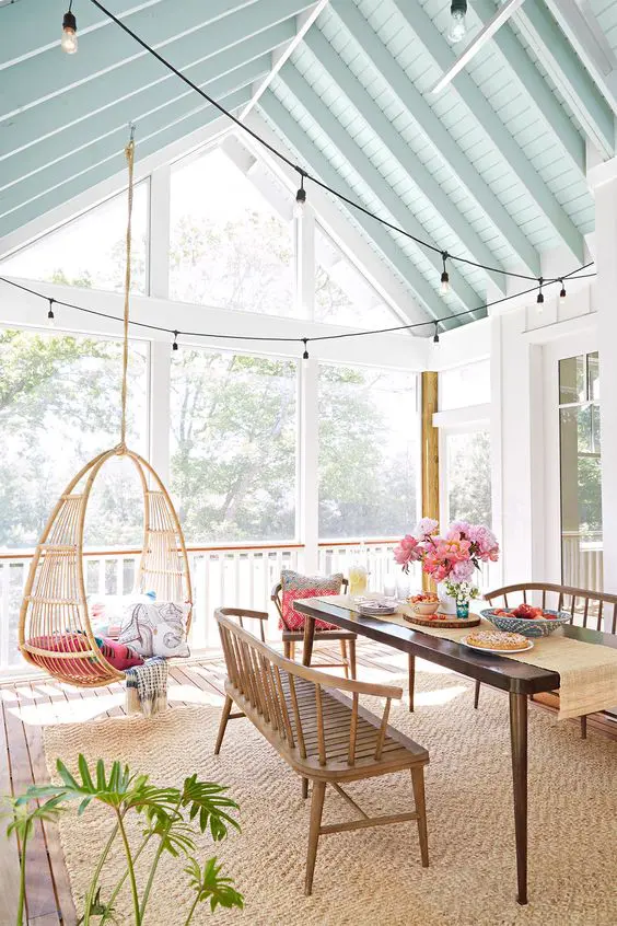a bright screened porch with a suspended chair, a stained table, benches and chairs, strign lights and greenery