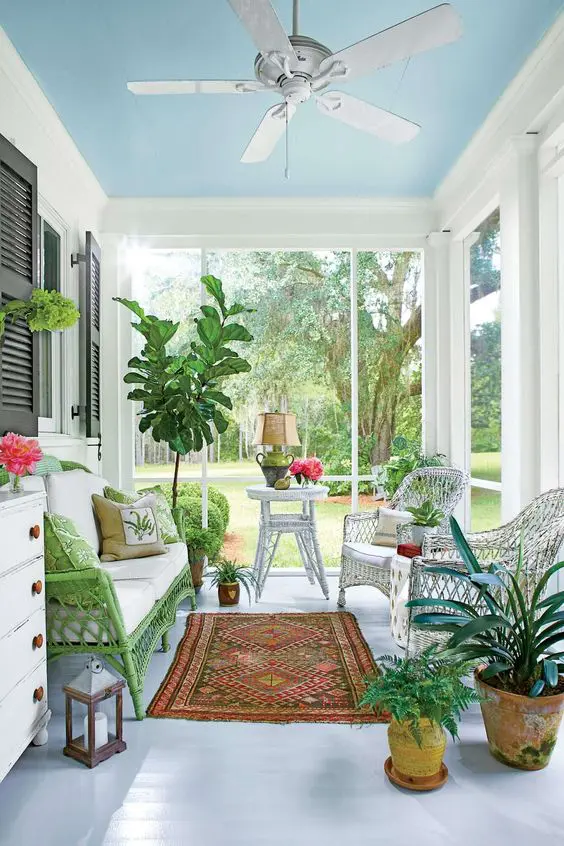 a colorful screened porch with a green woven loveseat, white chairs and side table, a white dresser, potted plants and flowers