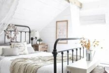a cozy farmhouse space with a forged bed in a niche, a white bench, some baskets and a wooden beam with lace curtains
