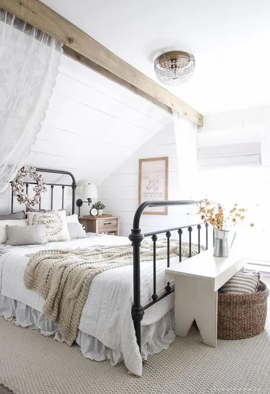 a cozy farmhouse space with a forged bed in a niche, a white bench, some baskets and a wooden beam with lace curtains