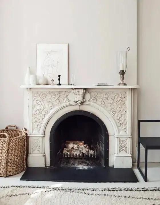 a delicately ornated French fireplace with a black chair and a basket next to it, some decor on the mantel and a printed rug