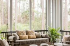 a greige screened porch with a suspended sofa, neutral chairs, side tables, a printed rug and potted ferns is a lovely space with a forest view