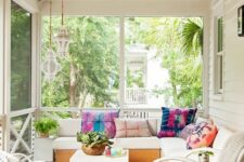 a lovely boho screened patio with a white corner sofa, white chairs, a coffee table and a leather pouf plus bold pillows and greenery