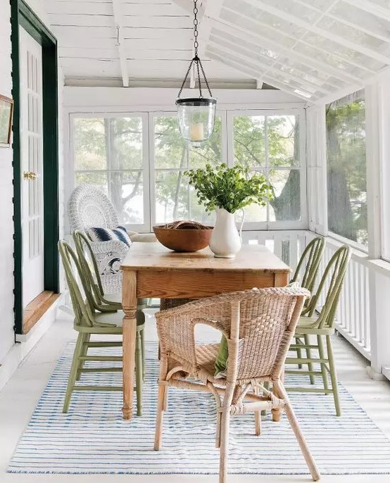 A lovely neutral screened porch with a stained table, green and neutral wicker chairs, some greenery   a cool dining space