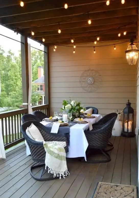 a lovely screened porch with a table and black woven chairs on comfortable stands, stirng lights, lanterns and greenery and blooms