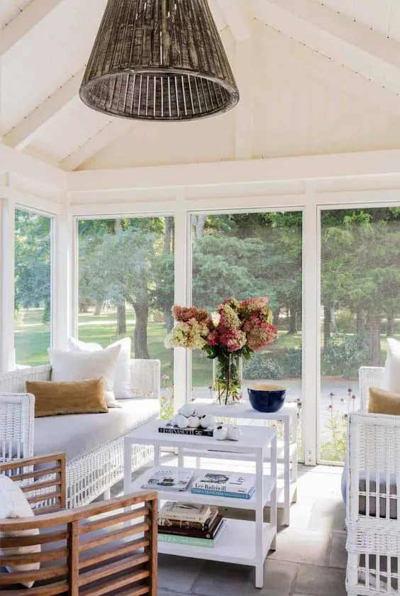 a lovely screened porch with white sofas, white tables and a curved chair plus blooms and a wicker pendant lamp