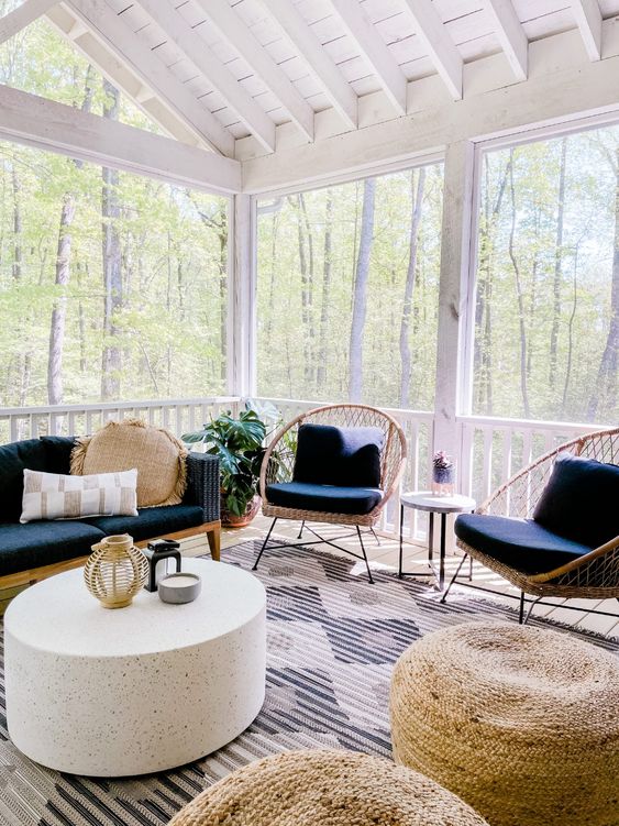 a modern boho screened porch with a navy sofa, rattan chairs, jute poufs, a coffee table and some potted greenery