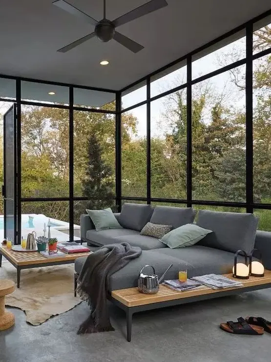 a modern screened patio with a grey corner sofa, low coffee tables and pillows and blankets plus a forest view