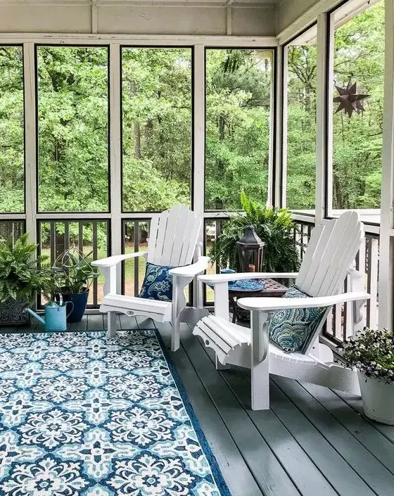 a modern screened patio with white wooden chairs, a blue printed rug and pillows, lots of potted greenery and greenery around