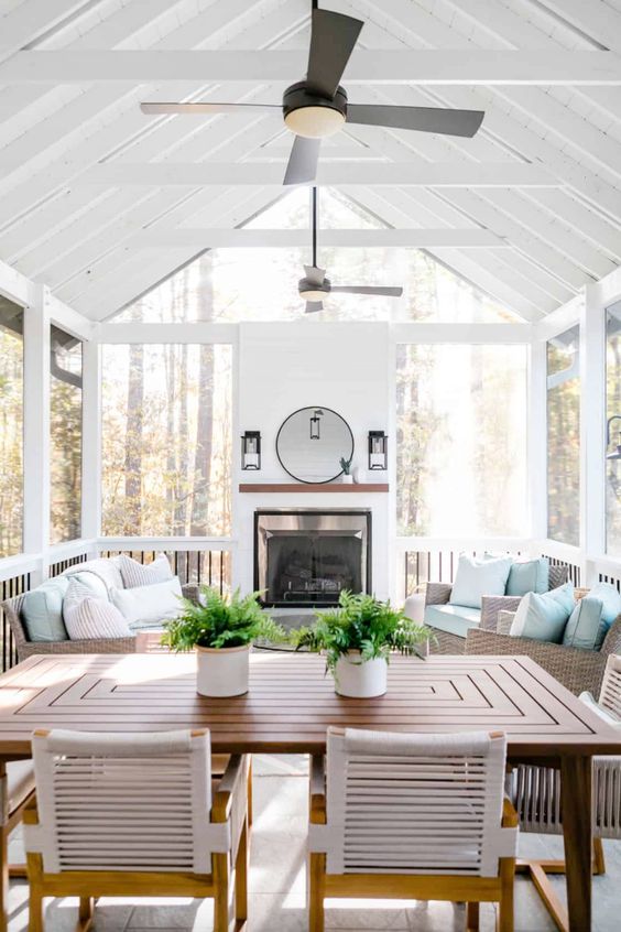 a screened farmhouse patio with a fireplace, wicker furniture with blue pillows, a stained dining set, potted greenery