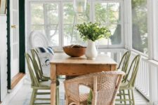 a screened porch with a stained table, green wooden and wicker chairs, a pendant lamp, some greenery and a striped rug