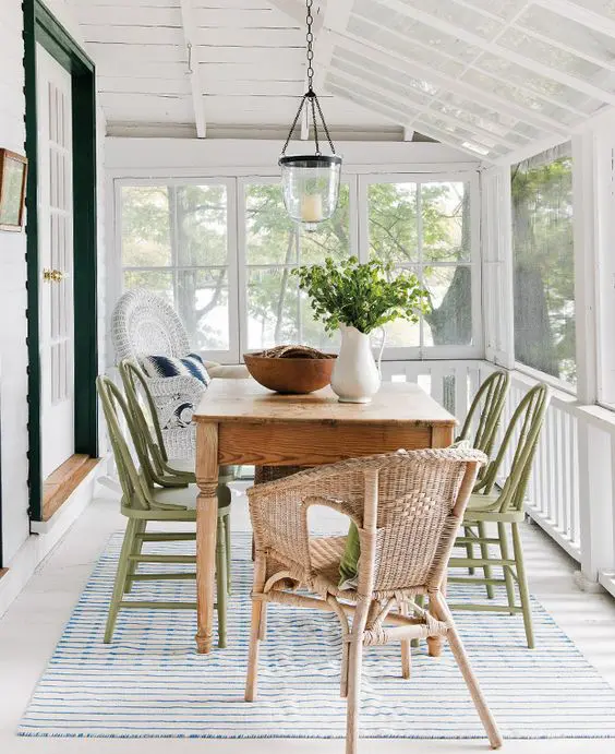a screened porch with a stained table, green wooden and wicker chairs, a pendant lamp, some greenery and a striped rug