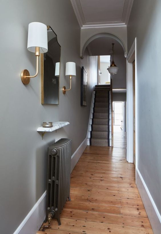 a small and elegant entryway with a dark radiator and a small shelf over it, with a mirror and wall lamps