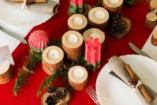 a Christmas tablescape with a red runner, red candles, wooden candleholders, pinecones, white plates, neutral napkins and cinnamon sticks