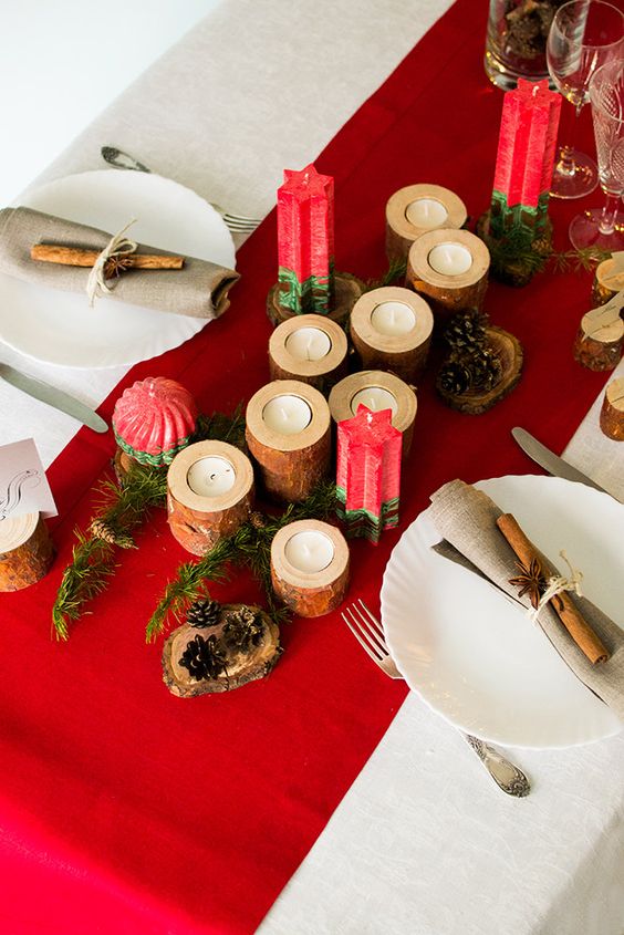 a Christmas tablescape with a red runner, red candles, wooden candleholders, pinecones, white plates, neutral napkins and cinnamon sticks