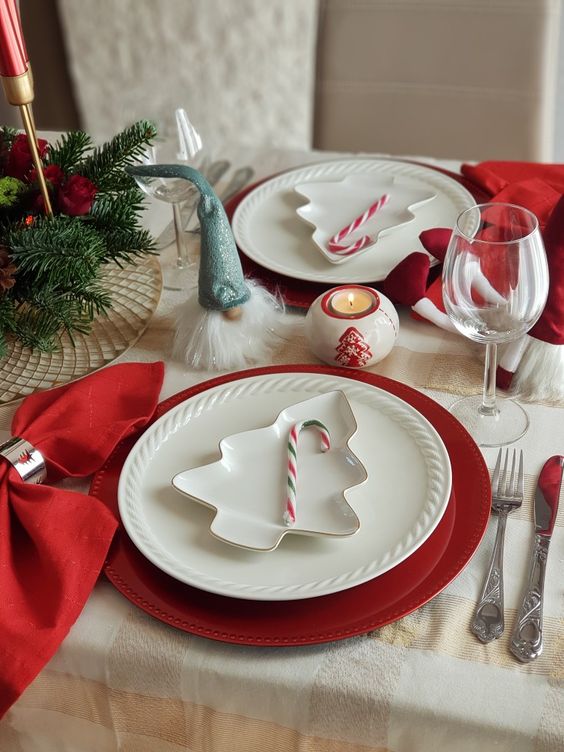 a Christmas tablescape with evergreens, red candles, napkins and chargers, white plates and candy canes