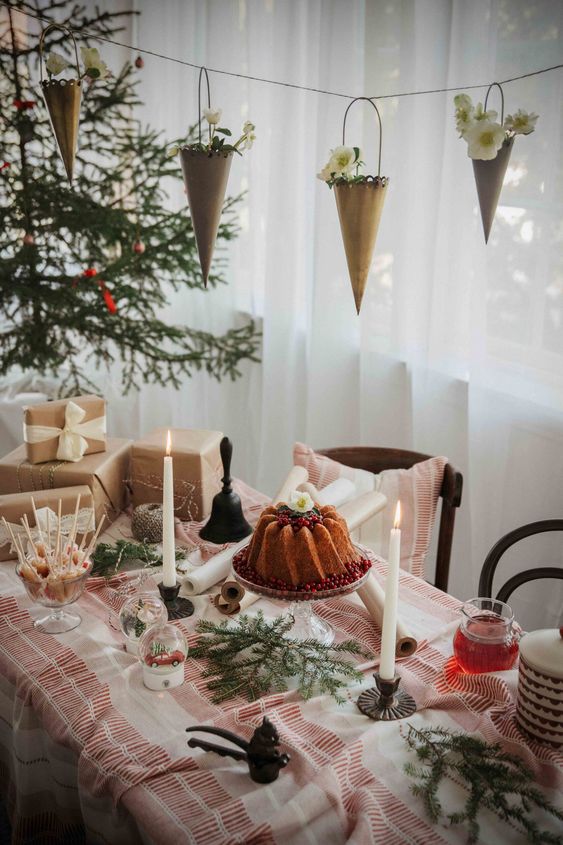 a Christmas tea party table with evergreens, a bundt cake, candles and sweets and printed textiles plus cones over the table
