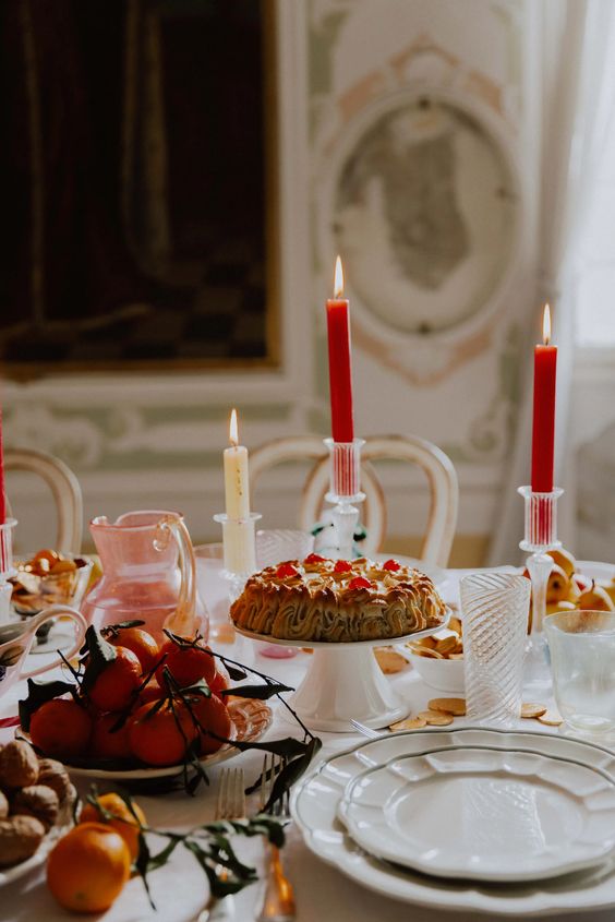 a beautiful Christmas tea party table with red candles, white porcelain, a cake, some fruit and colored jugs and glasses