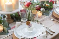a cozy Christmas tea party table with pillar candles on stands, greeneyr and pomegranates, wood slice placemats, white porcelain and sweets