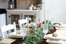 a pretty Christmas tablescape with an evergreen and berry runner, candles, white porcelain and gingerbread houses on each plate