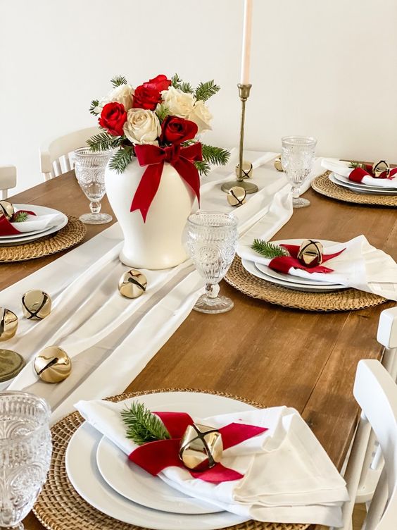 a simple and elegant Christmas table setting with white textiles, red and white roses, bells, woven placemats
