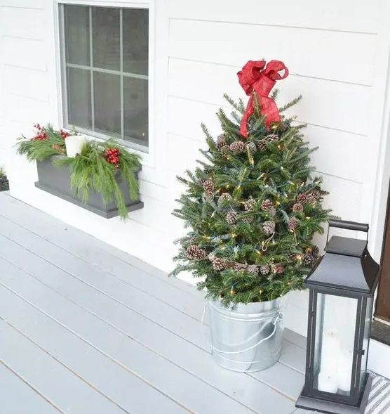 a small holiday tree decorated with lights, pinecones and a red bow on top in a galvanized bucket for a porch