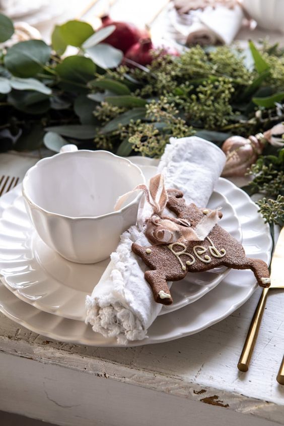 a stylish Christmas tea party table with white porcelain, a greenery and pomegranate runner, a gingerbread cookie and elegant cutlery