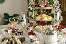 an elegant Christmas tea party table with a tablecloth, tiered stands with sweets and cookies, printed teaware and candles and evergreens