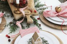 an eye-catchy Christmas tea party table with a centerpiece of candles and candle lanterns with cranberries, wooden placemats, plates with bells and gingerbread houses