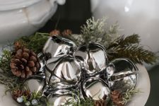 a Christmas arrangement of a white bowl, evergreens, pinecones and silver bells is a lovely decoration for the holidays