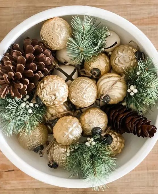 A Christmas centerpiece of a white bowl with bells, gold ornaments, pinecones and evergreens is a cool vintage inspired decor idea