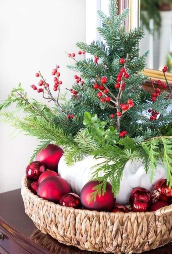 a Christmas centerpiece of a woven bowl, red ornaments, a teapot with evergreens and berries for a rustic space