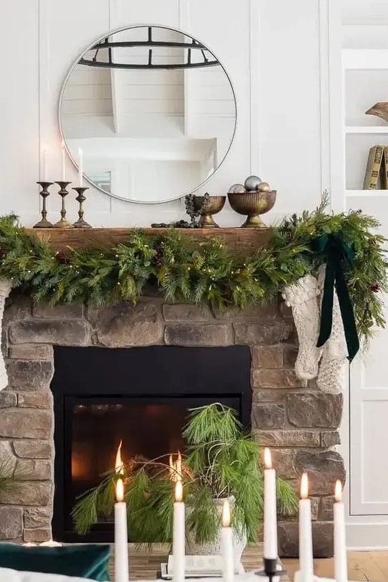 a Christmas mantel with an evergreen and light garland with pinecones, white stockings and a green bow, candles in candleholders