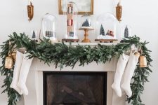 a Christmas mantel with an evergreen garland, bells, stockings, cloches with mini trees and houses