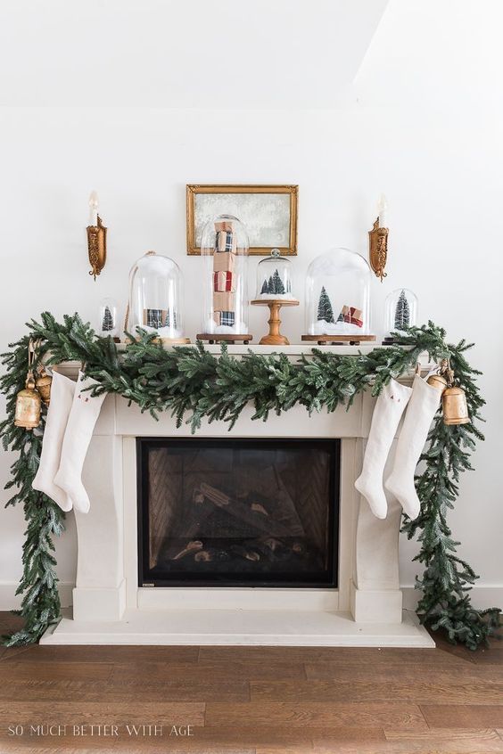 a Christmas mantel with an evergreen garland, bells, stockings, cloches with mini trees and houses