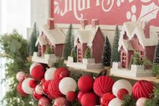 a catchy Christmas mantel decorated with evergreens, bold red and white ornaments, pink houses and bottle brush Christmas trees
