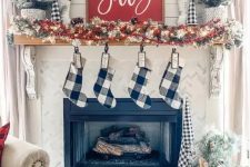 a cozy farmhouse christmas mantel with a snowy pinecone and light garland, red berries, snowy mini trees and buffalo check stockings