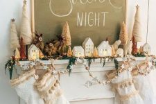 a holiday mantel with white tasseled stockings, an evergreen and light garland, stars and wooden beads, small houses and Christmas trees