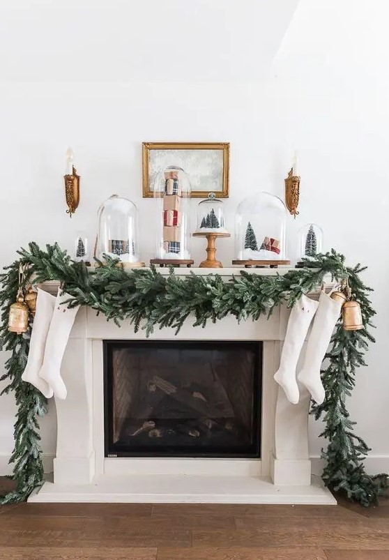 a lovely Christmas mantel with a lush evergreen garland, cloches with mini trees and gift boxes, oversized balls and stockings