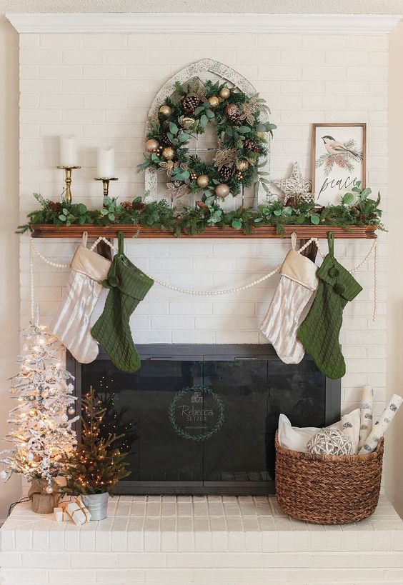 a lovely Christmas mantel with an evergreen garland and wreath, white and green stockings, mini trees and candles