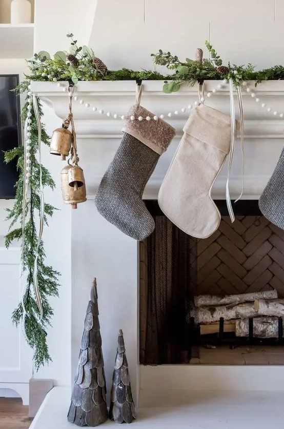 a modern Christmas mantel with a greenery and pinecone garland, bells, stockings and a pompom garland for a touch of fun