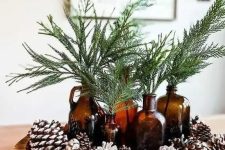 a natural woodland Christmas centerpiece of a wooden bowl with pinecones, evergreens in dark glass bottles is cool