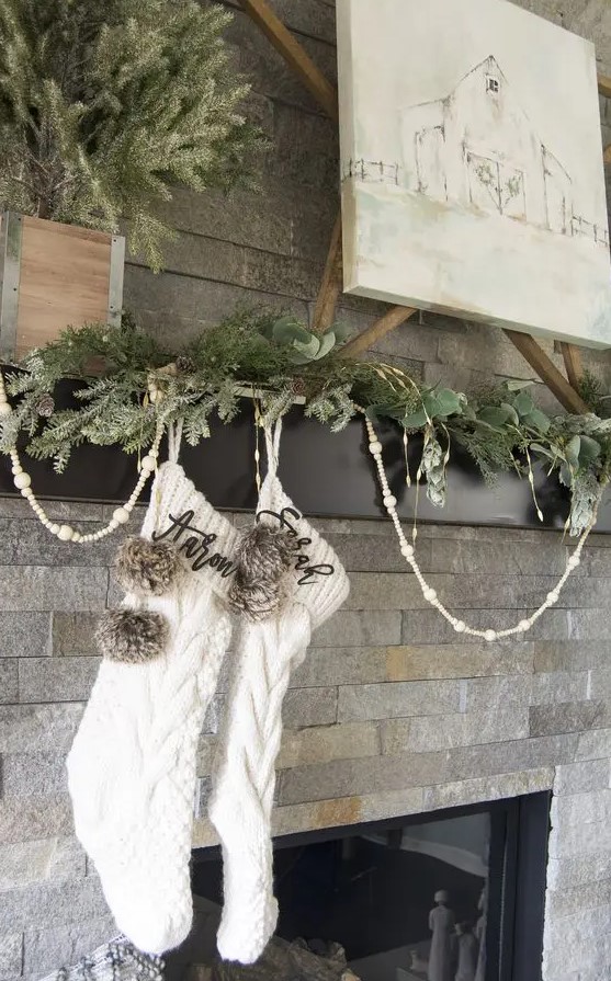 a pretty modern farmhouse mantel with an evergreen garland, wooden beads, white stockings and pompoms is amazing
