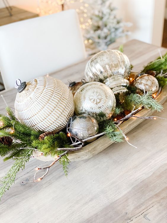 a rustic Christmas centerpiece of a dough bowl with evergreens, pinecones, mercury glass ornaments, twigs and lights