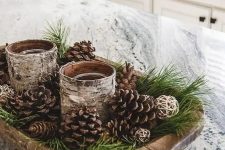 a rustic Christmas centerpiece of a wooden bowl with evergreens, pinecones and candles wrapped with bark